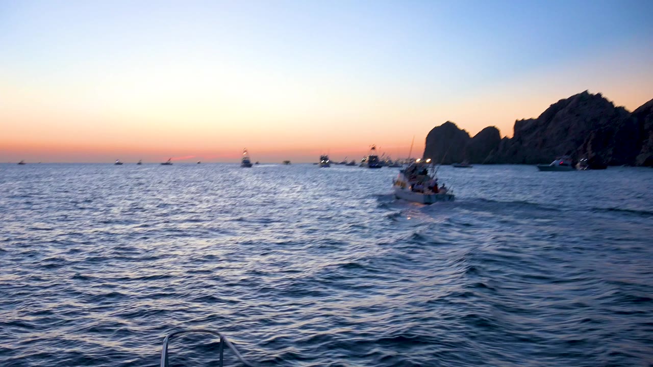 Cabo San Lucas sunset scene with the small boat farther right, a broad field of rippled water, clustered boats near the horizon, dark rocks on the right, and no visible people.