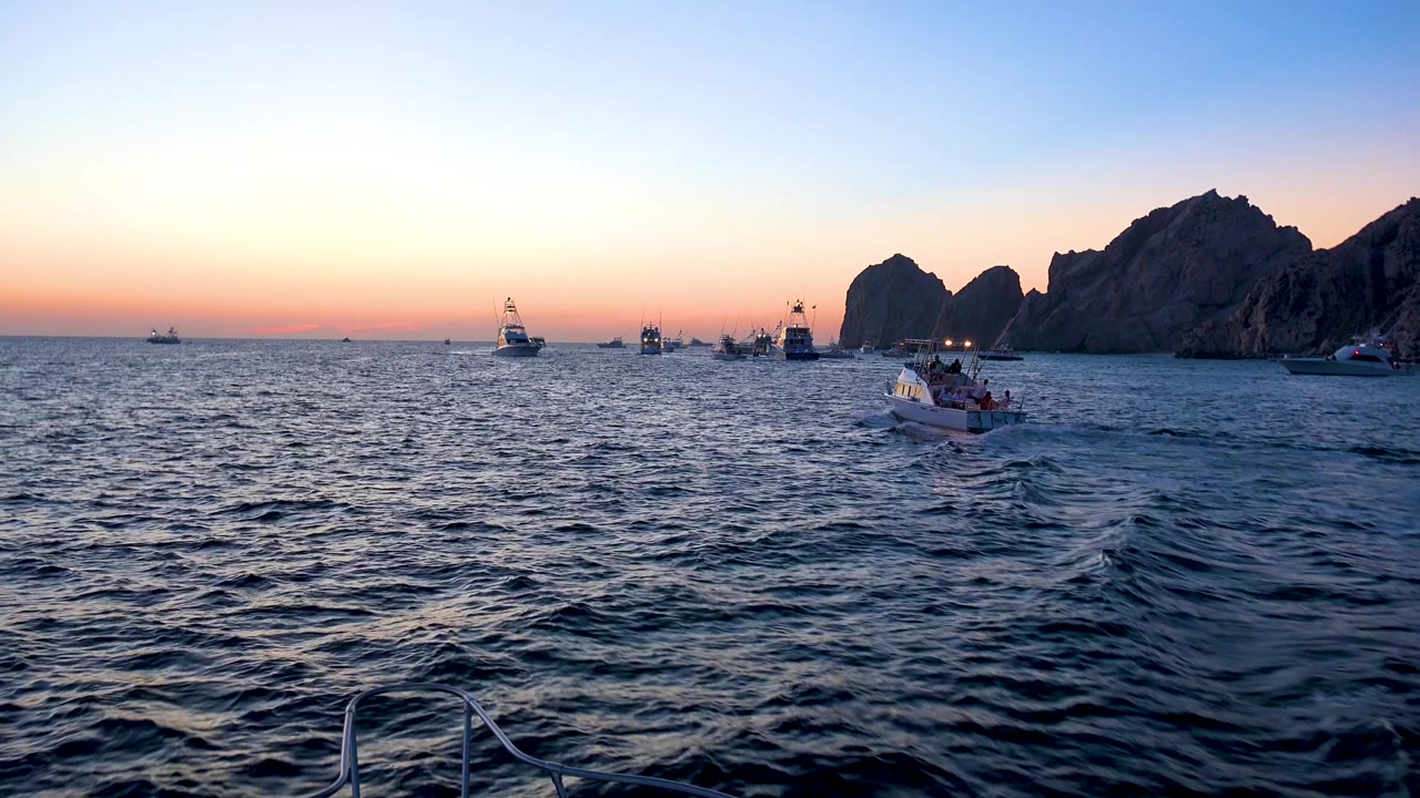 Cabo San Lucas evening water view with multiple small boats spaced across the midground, silhouetted rocks on the right, a peach horizon, and no visible people.