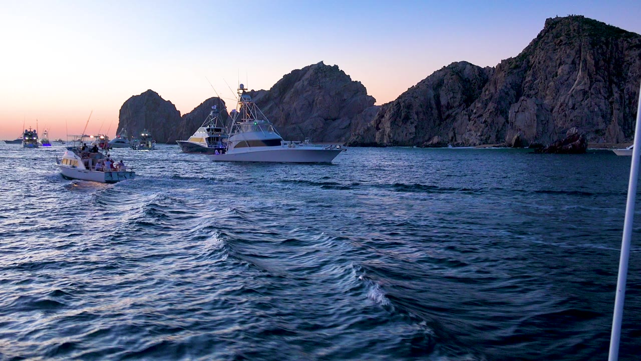 Cabo San Lucas, Mexico frame showing a small boat left of center and a white sportfishing boat near rocky mountains, with textured blue water and no clearly visible people.