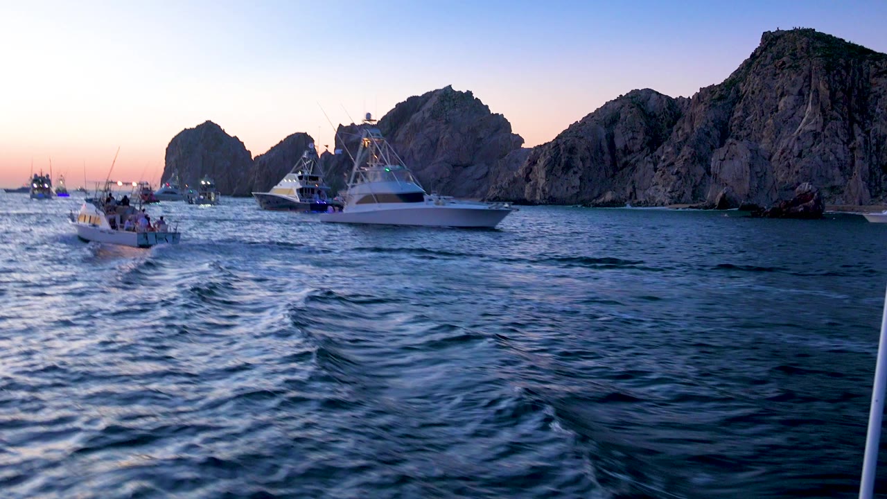 Cabo San Lucas, Mexico frame with a white sportfishing boat right of center, smaller boats to the left, choppy foreground water, rocky mountains, and no clearly visible people.