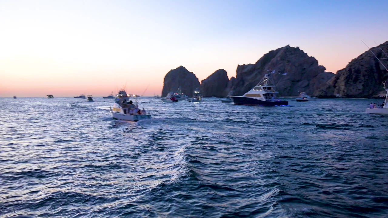 Multiple boats spread across the water near Cabo San Lucas rock formations, with a large sportfishing boat on the right, textured waves in front, and no visible people.