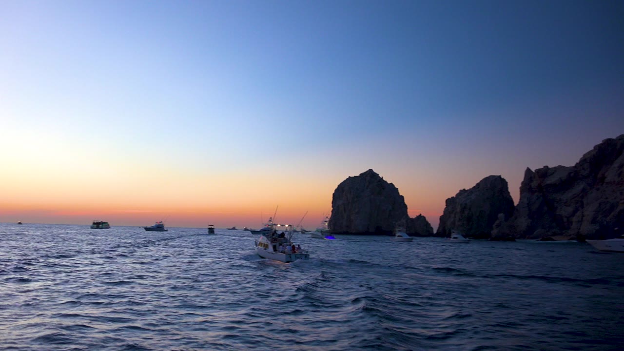 Cabo San Lucas twilight ocean view with several small boats on the horizon, a tour boat near center-right, dark rock formations at right, and no visible people.