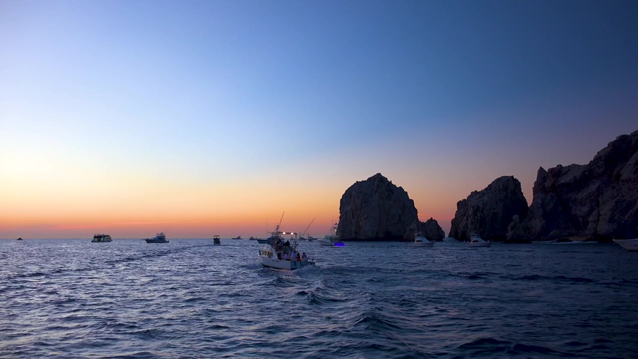 Cabo San Lucas dusk seascape with a low horizon, scattered boats across the water, a midground boat wake, shaded rock formations on the right, and no visible people.