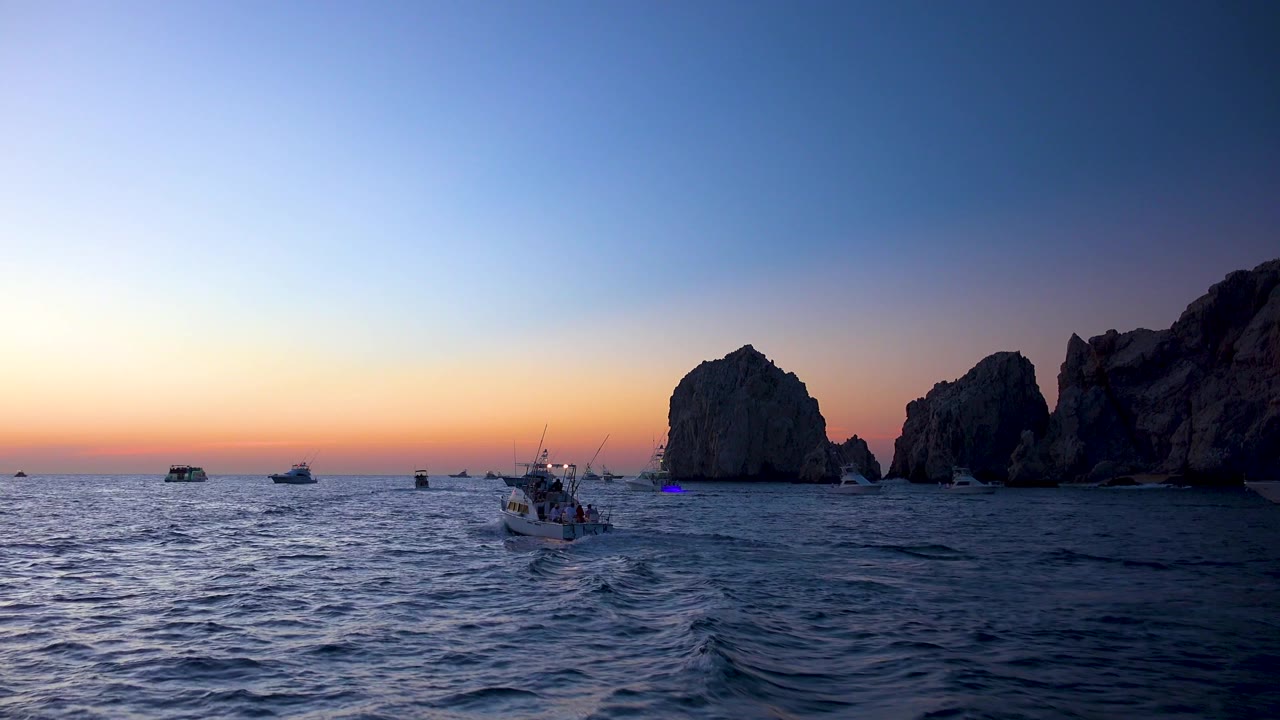 Cabo San Lucas open-water sunset view with darker waves in the foreground, a tour boat wake near center, distant boats to the left, rock formations to the right, and no visible people.