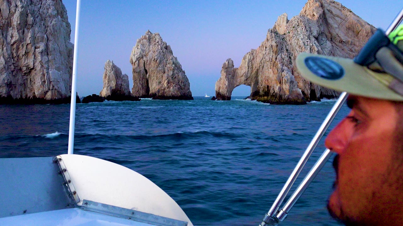 Boat rail and a partial passenger face frame blue water and the Cabo San Lucas rock arch in bright daylight.