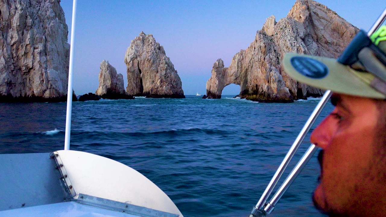 A partial passenger profile and boat hardware sit close to camera while the Cabo San Lucas arch and sea stacks remain across the water.