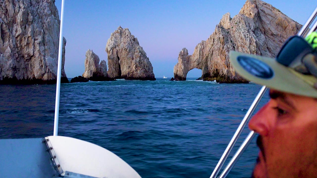 The rocky arch and adjacent sea stacks are visible beyond dark blue water, with a cropped passenger face and boat railing at the right edge in Cabo San Lucas.
