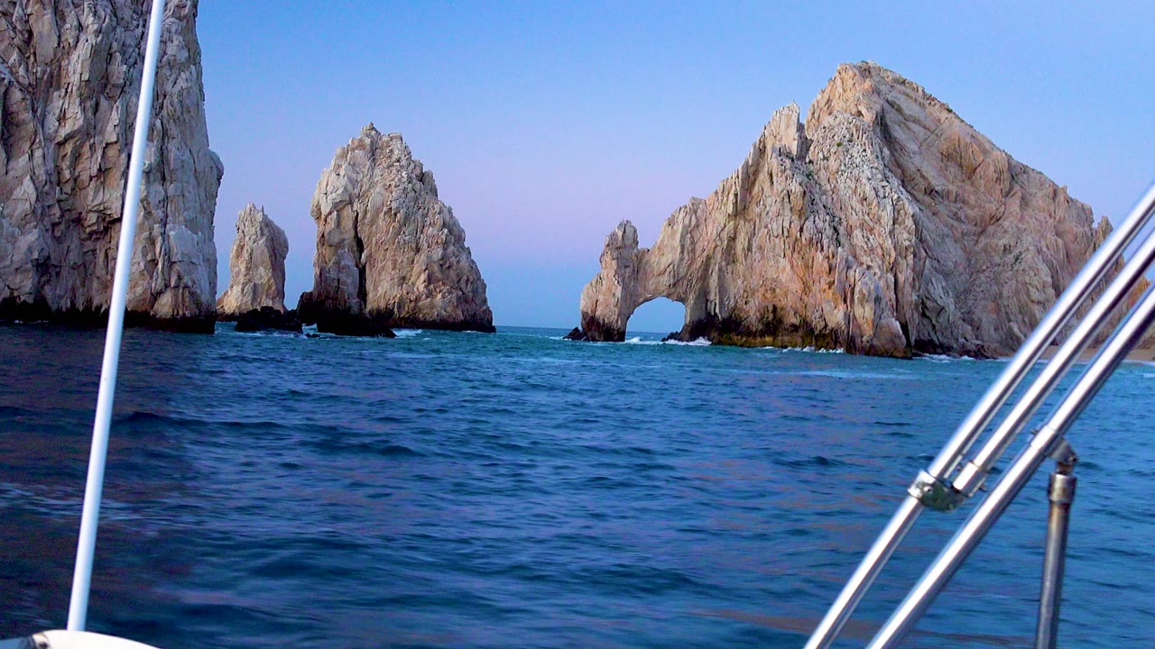 Cabo San Lucas rock formations and a natural arch rise above blue ocean, viewed from a boat with metal railings in the foreground and no visible people.