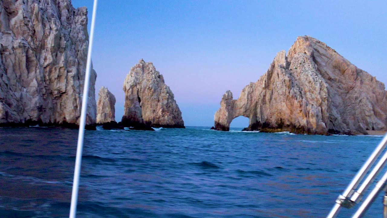 A slightly tilted Cabo San Lucas boat view shows blue water, pale rock formations, a natural arch, a cropped cliff edge at left, and no visible people.