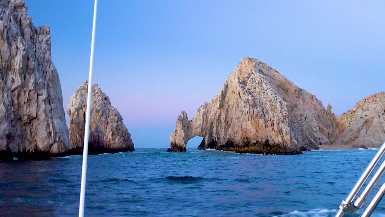 Cabo San Lucas rock formations sit across the horizon as blue water fills the foreground and a boat rail enters from the lower right, with no visible people.