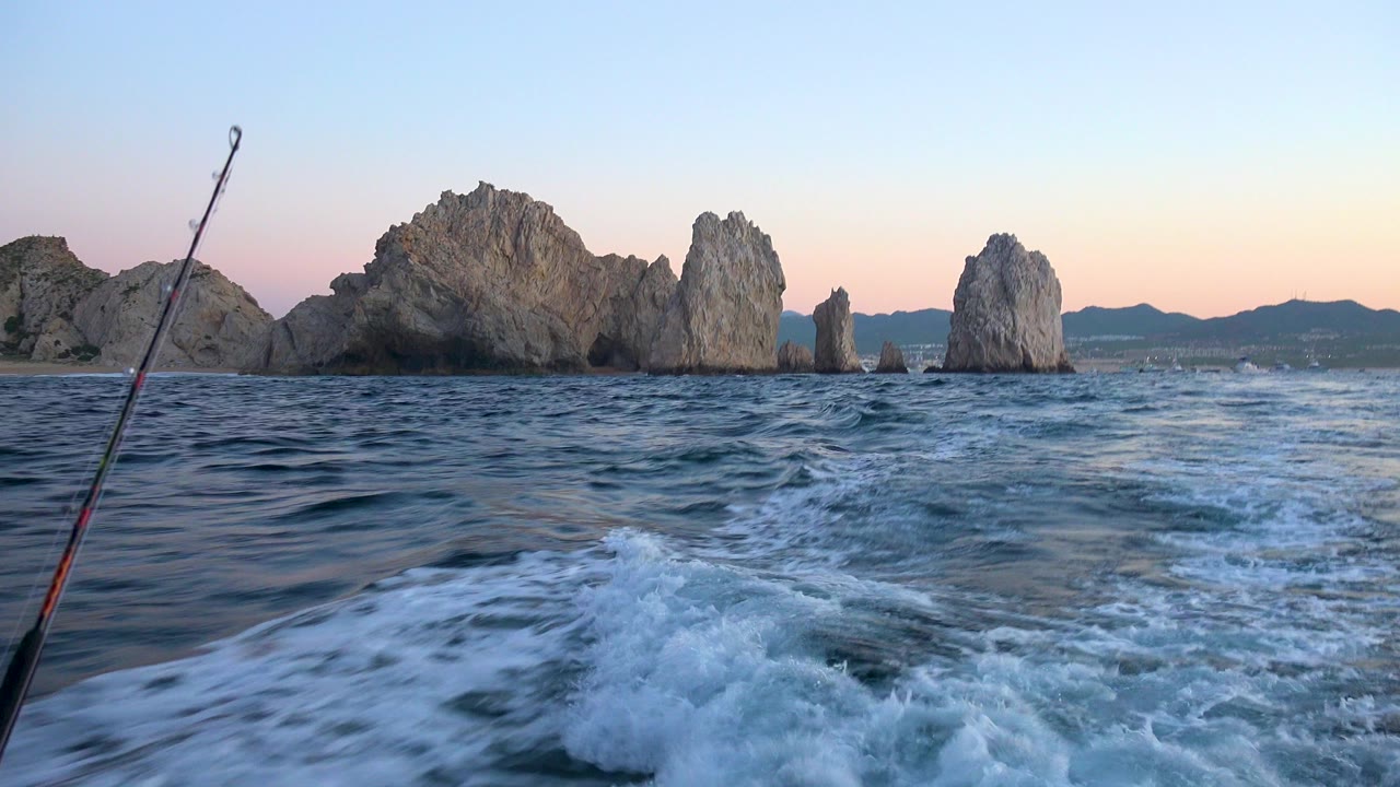 Cabo San Lucas ocean view from a boat with a fishing rod at the left edge, foamy wake in the foreground, jagged rock formations ahead, and no visible people.