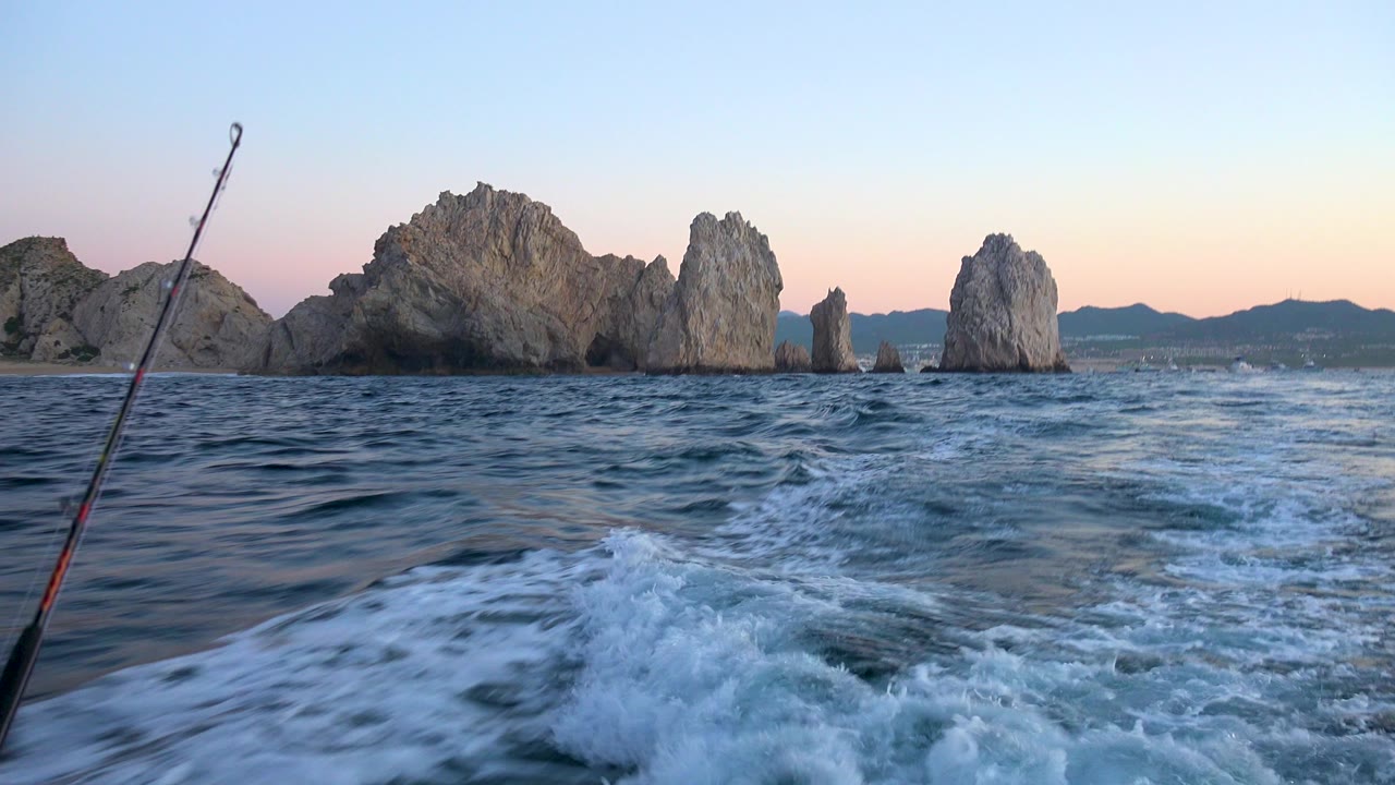 Cabo San Lucas water scene with a fishing rod leaning into frame, white wake spreading across the lower image, clustered rock formations beyond, and no visible people.