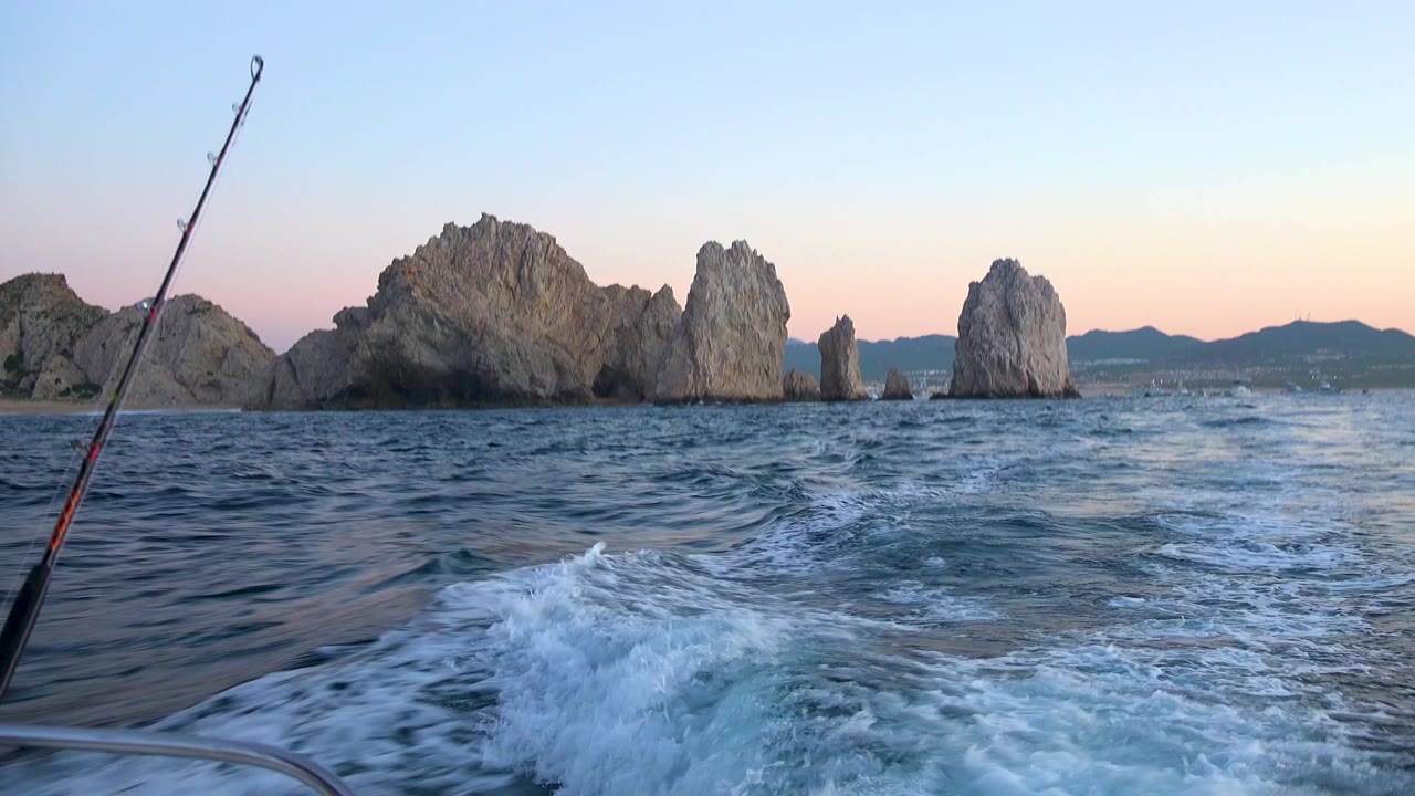 Cabo San Lucas sunset frame with a fishing rod near the left side, curved boat wake in the foreground, rocky sea stacks across the middle, distant boats, and no visible people.