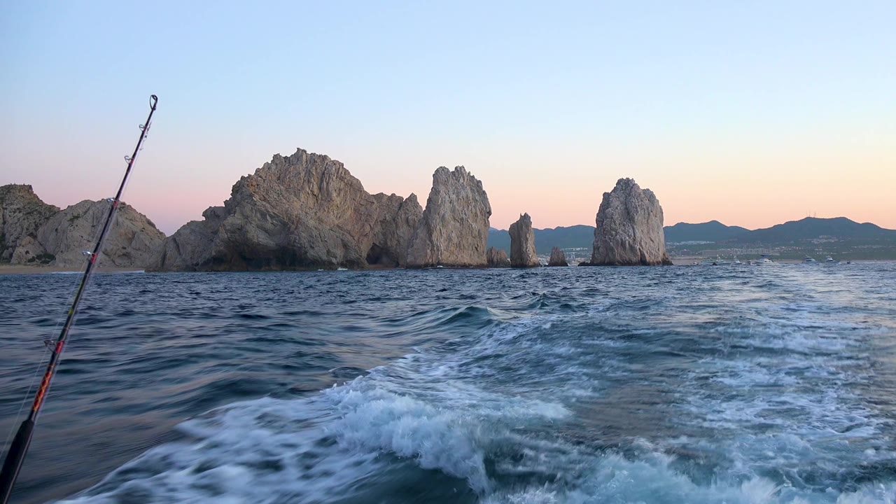Cabo San Lucas ocean view with the fishing rod along the left border, smoother wake in the foreground, large gray rock formations centered, mountains on the horizon, and no visible people.