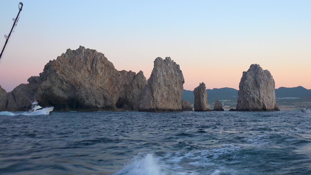 Cabo San Lucas rock stacks fill the center of the frame while blue ocean chop and white wake move across the foreground, with no visible people.