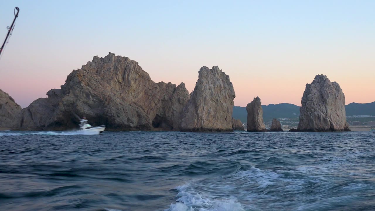 A small boat sits near breaking water at the base of Cabo San Lucas rock formations, with rippled ocean in front and no visible people.