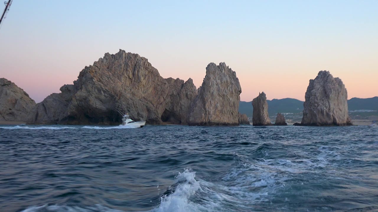 White boat wake cuts through dark blue water toward Cabo San Lucas rock formations and distant hills beneath a pale sunset sky, with no visible people.