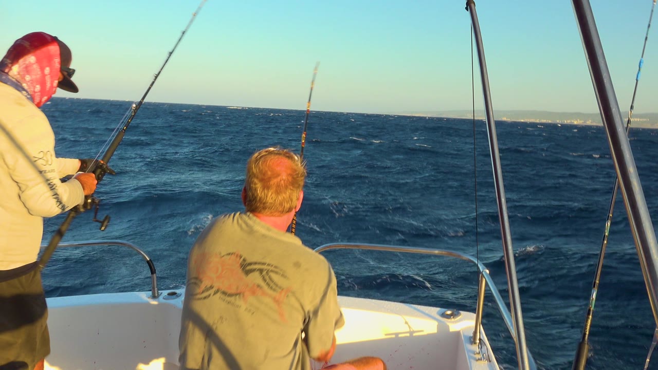 Two men sit and stand near the bow of a small boat off Cabo San Lucas, Mexico, with fishing rods angled over choppy blue water in warm sunlight.