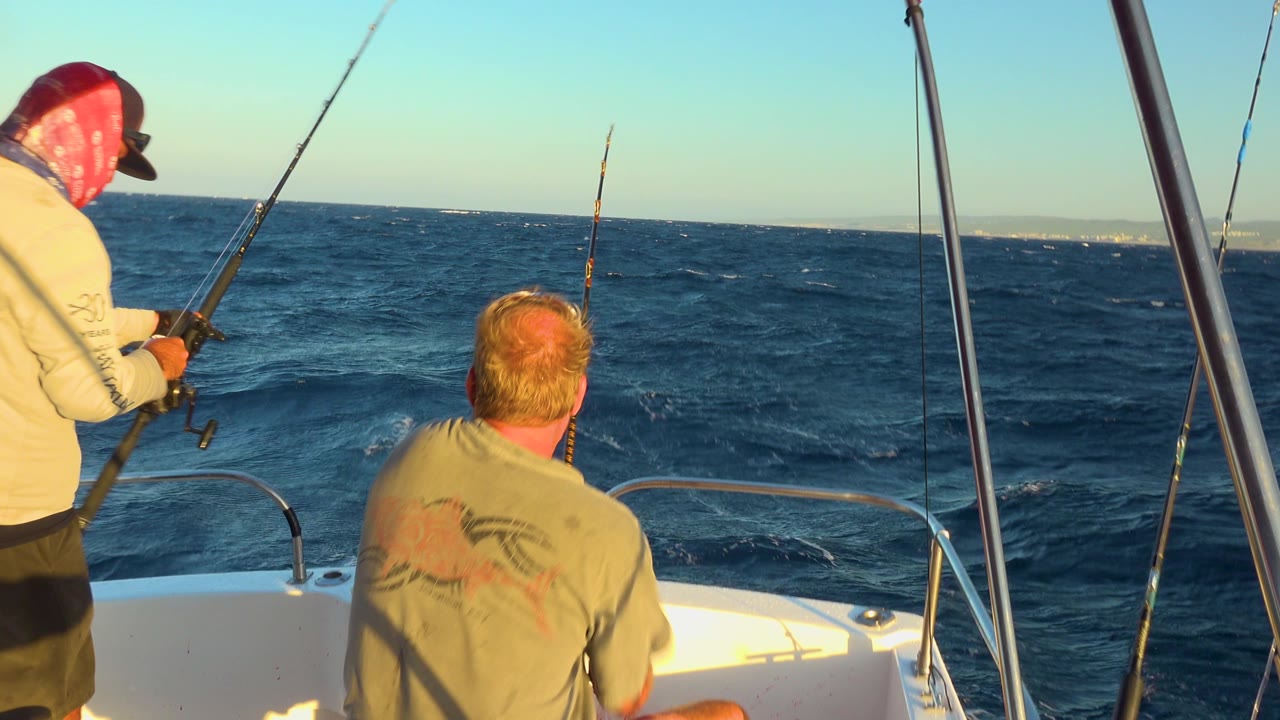 Two men on a boat watch fishing lines over dark blue water.