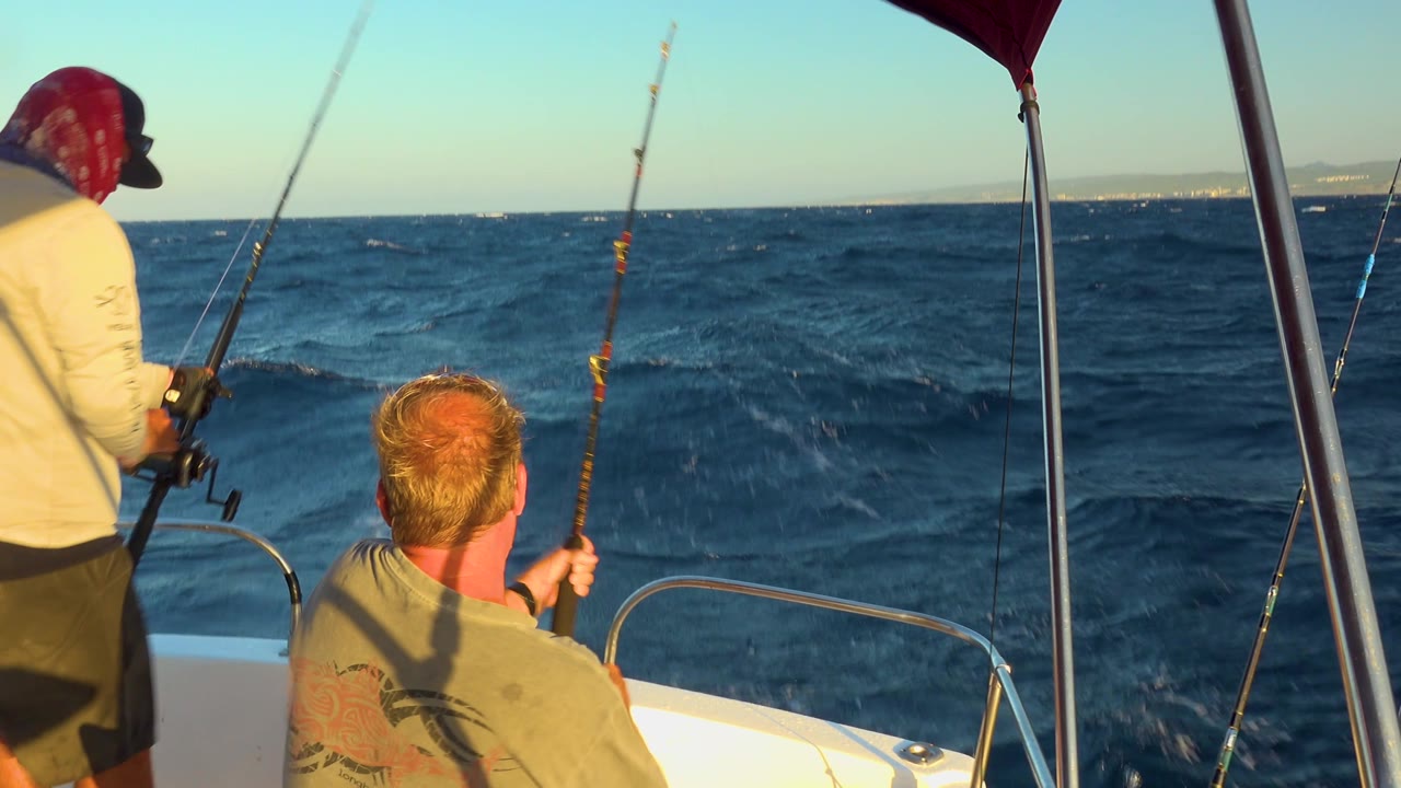 A seated man raises his fishing rod while a second man in a red head covering stands to the left, with sunlit waves behind the bow of the boat in Cabo San Lucas.