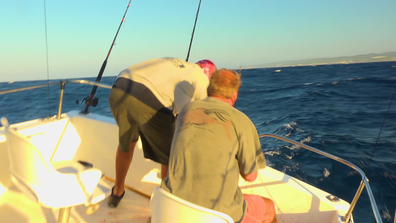 Two men on a small boat off Cabo San Lucas, Mexico lean toward the bow while fishing rods stand against choppy blue water.