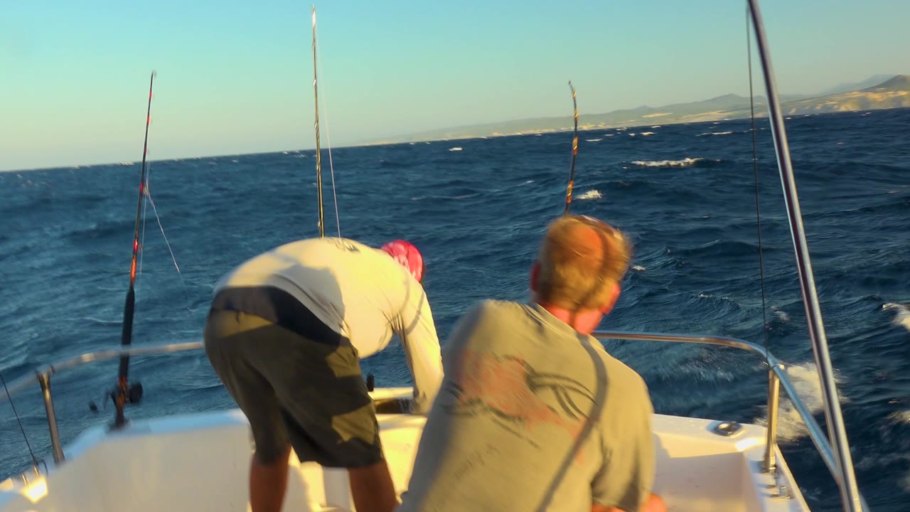 A man in a light shirt bends over the bow while another man sits nearby, with multiple fishing rods and rolling Cabo San Lucas water behind them.