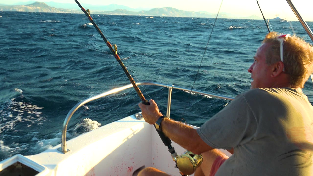 At 0:00 in Cabo San Lucas, Mexico, an adult man grips a fishing rod near the bow rail while dark blue water and distant mountains fill the background.