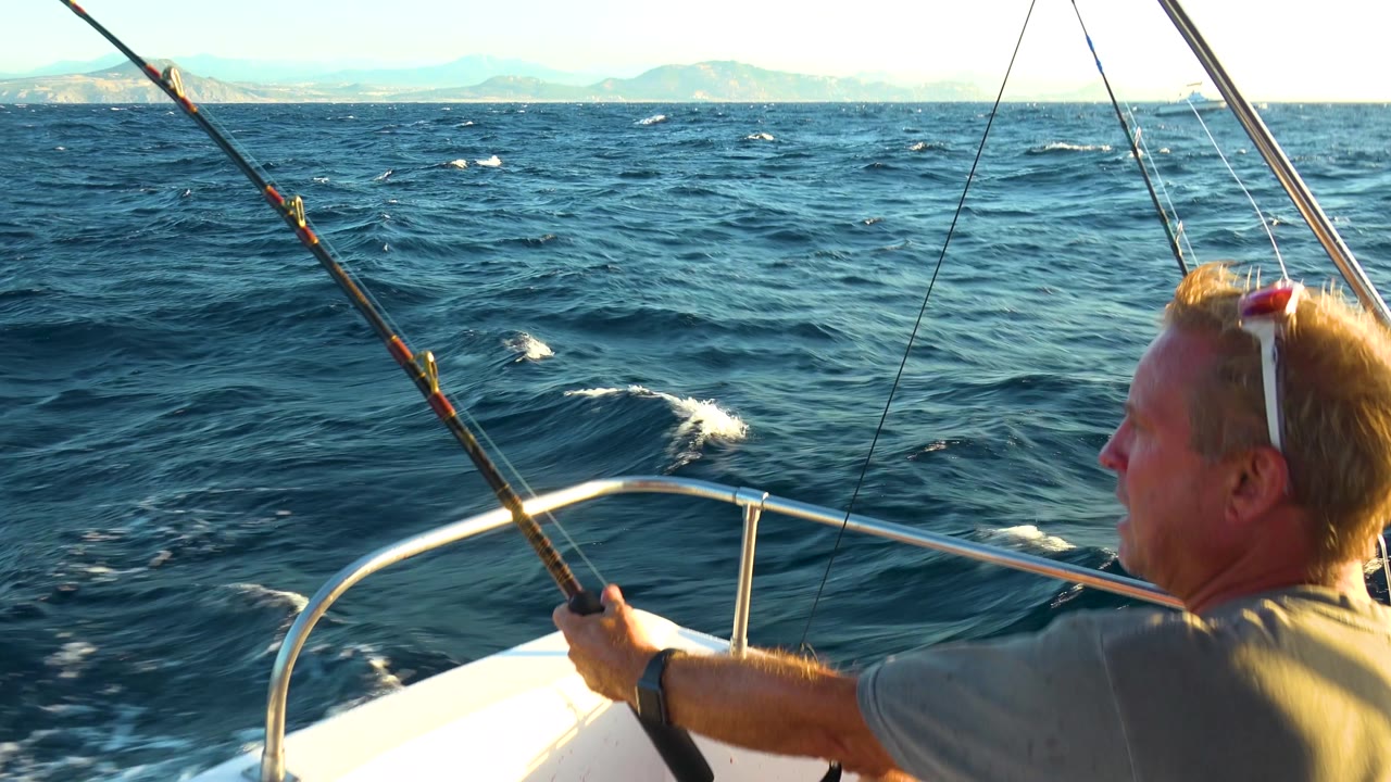 At 0:02 in Cabo San Lucas, Mexico, the fisherman is partly cropped on the right while the rod, bow rail, rolling waves, and pale mountain horizon dominate the frame.
