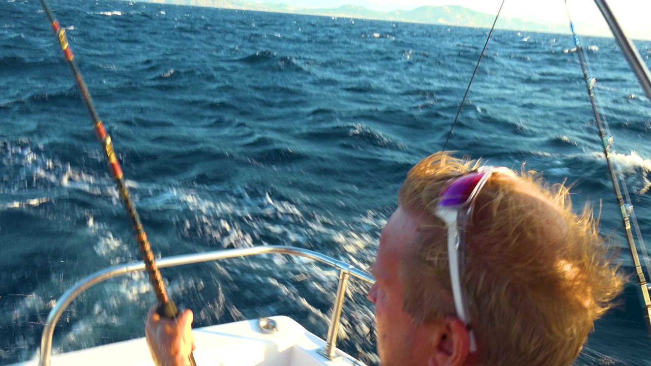 A blond man with sunglasses on his head holds a fishing rod near the bow rail of a boat in Cabo San Lucas, Mexico, with choppy blue sea and distant mountains behind him.