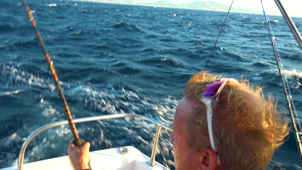 Man on a boat holds a fishing rod as waves pass beside the bow.