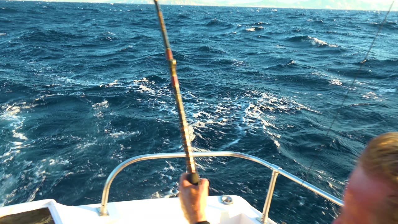 A fishing rod rises from the boat bow in Cabo San Lucas, Mexico, while choppy water dominates the frame and the fisherman's head is partly cropped on the right.