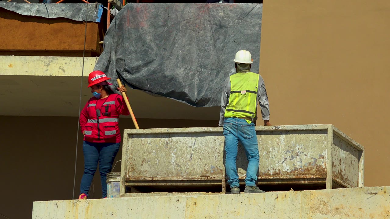 At 0:00 in Cabo San Lucas, Mexico, a woman in a red jacket and hard hat stands near a second worker in a yellow safety vest beside a weathered metal bin on a concrete level.