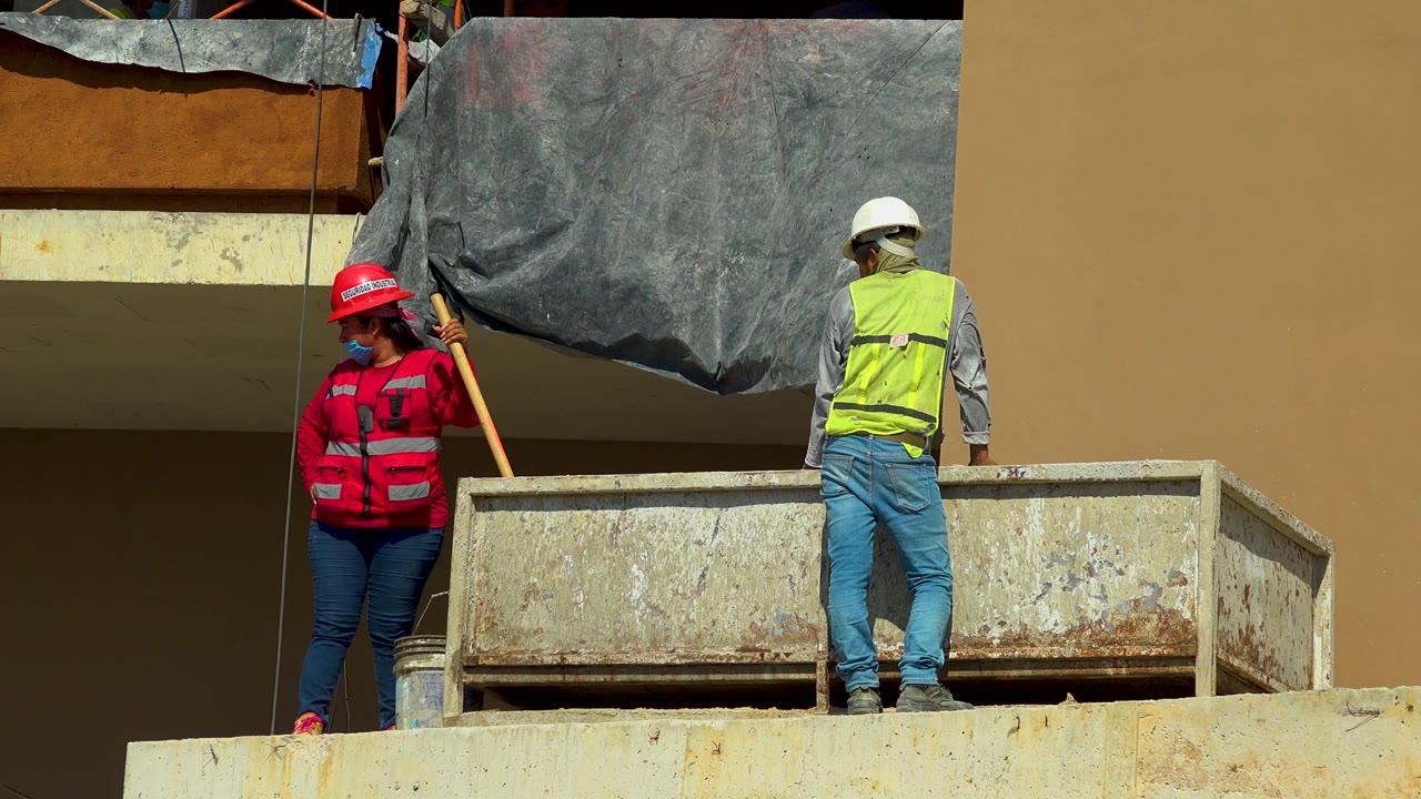 At 0:02 in Cabo San Lucas, Mexico, the woman in red stands near a vertical pole while the other worker faces the building, with a black tarp draped above the metal bin.