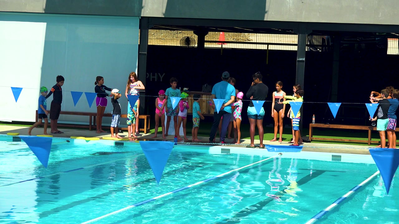 Adults and children stand along a turquoise pool in Cabo San Lucas, Mexico, with blue triangular lane flags crossing the foreground and a shaded wall behind them.