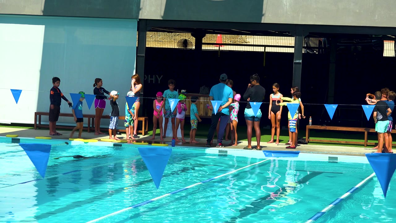 People in swimwear cluster at the deck in Cabo San Lucas, Mexico, while blue lane flags run across the lower foreground over the bright pool surface.
