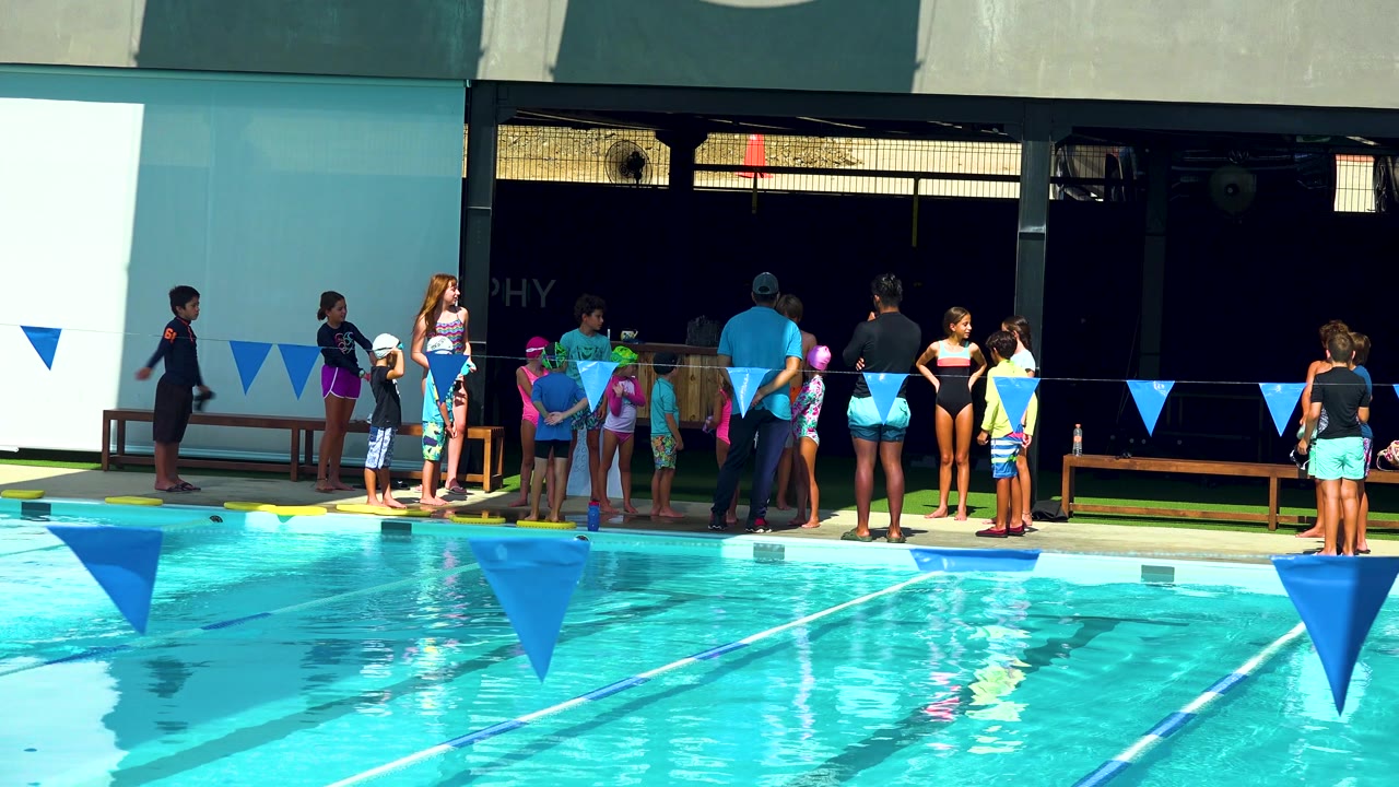 In Cabo San Lucas, a group of children in bright swimsuits waits near adults on the pool deck while blue lane flags cross the frame above the water.