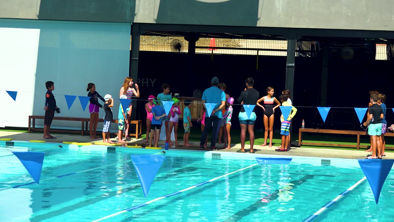 In Cabo San Lucas, swimmers remain gathered on the deck, with turquoise pool water in the foreground and a dark shaded wall behind the group.