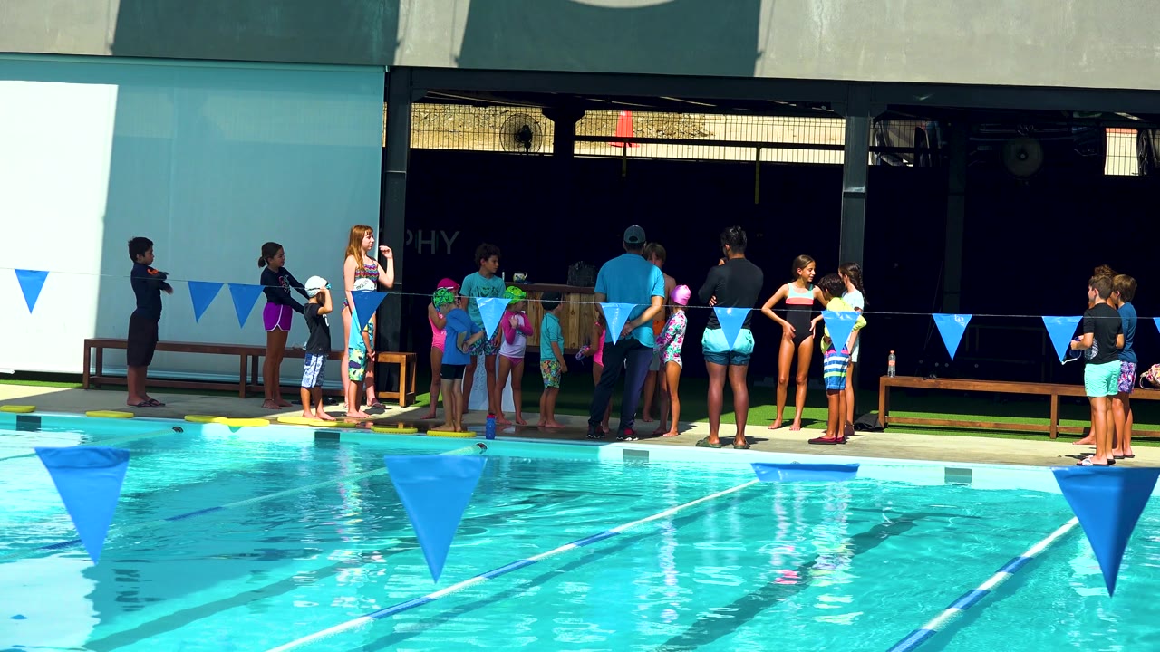 Adults and children stand on a sunny pool deck beside lane flags.