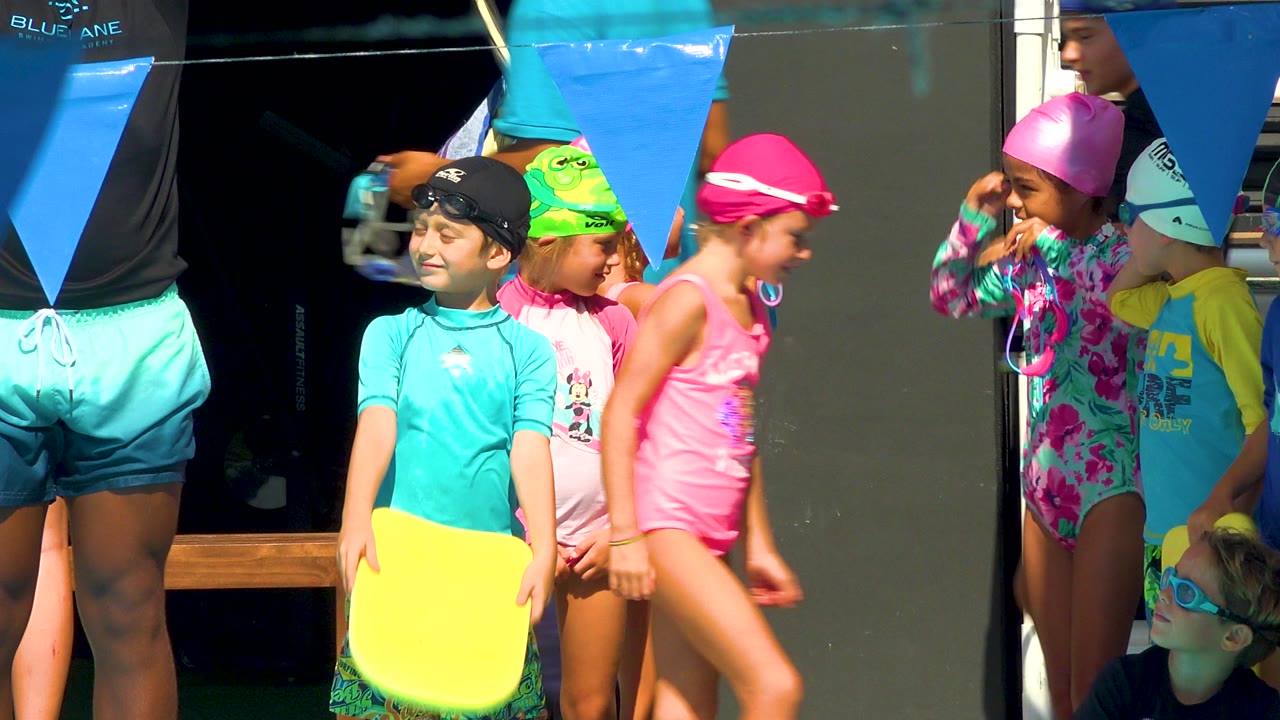 Children in colorful swim caps and swimsuits stand poolside in Cabo San Lucas while a yellow kickboard and blue pennant flags fill the foreground.