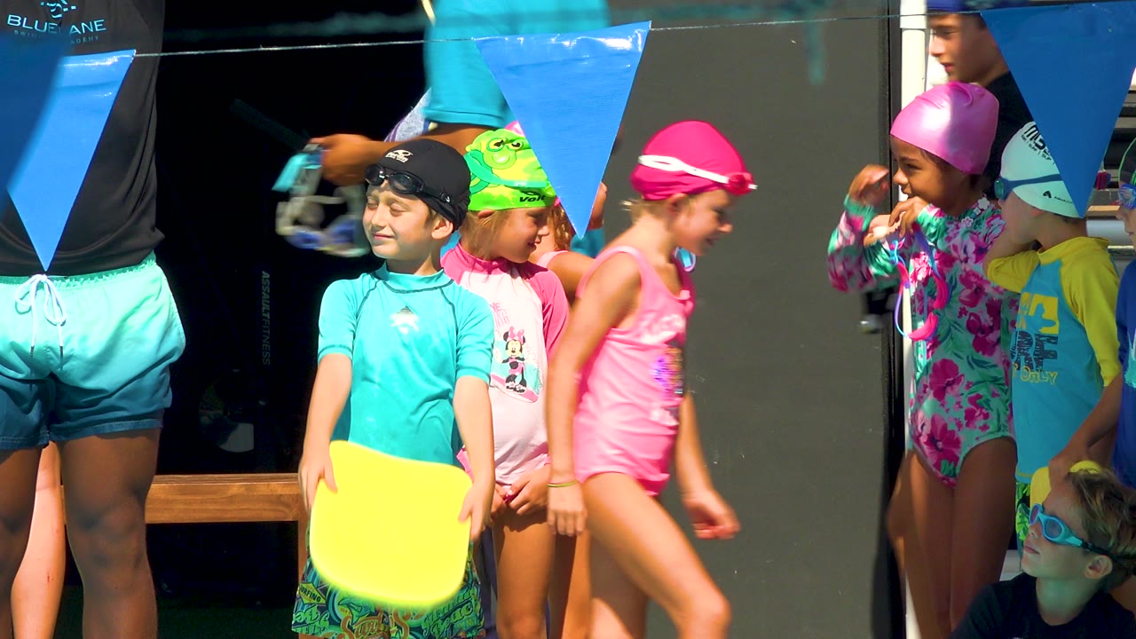 A child in a turquoise swim shirt holds a yellow kickboard at chest height while other children in pink, green, and purple caps gather beside blue pennant flags in Cabo San Lucas.