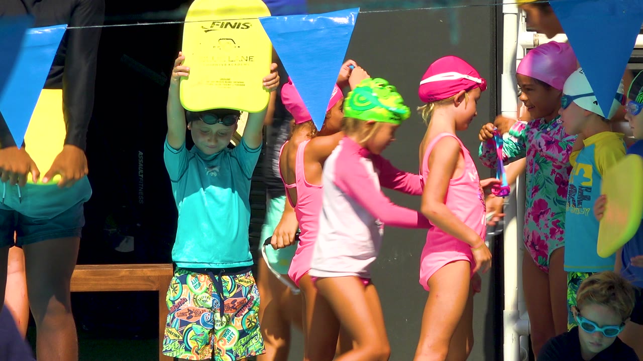 A child raises a yellow kickboard overhead in Cabo San Lucas as children in pink swimsuits and bright swim caps pass and stand beneath blue triangular pennants.