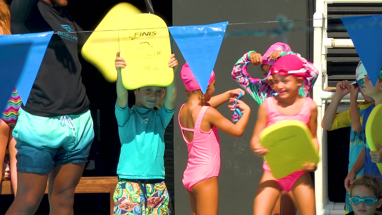 At the start of the Cabo San Lucas clip, children in bright swim caps and goggles stand near blue pennant flags while holding yellow swim boards, with an adult partly visible at the edge.