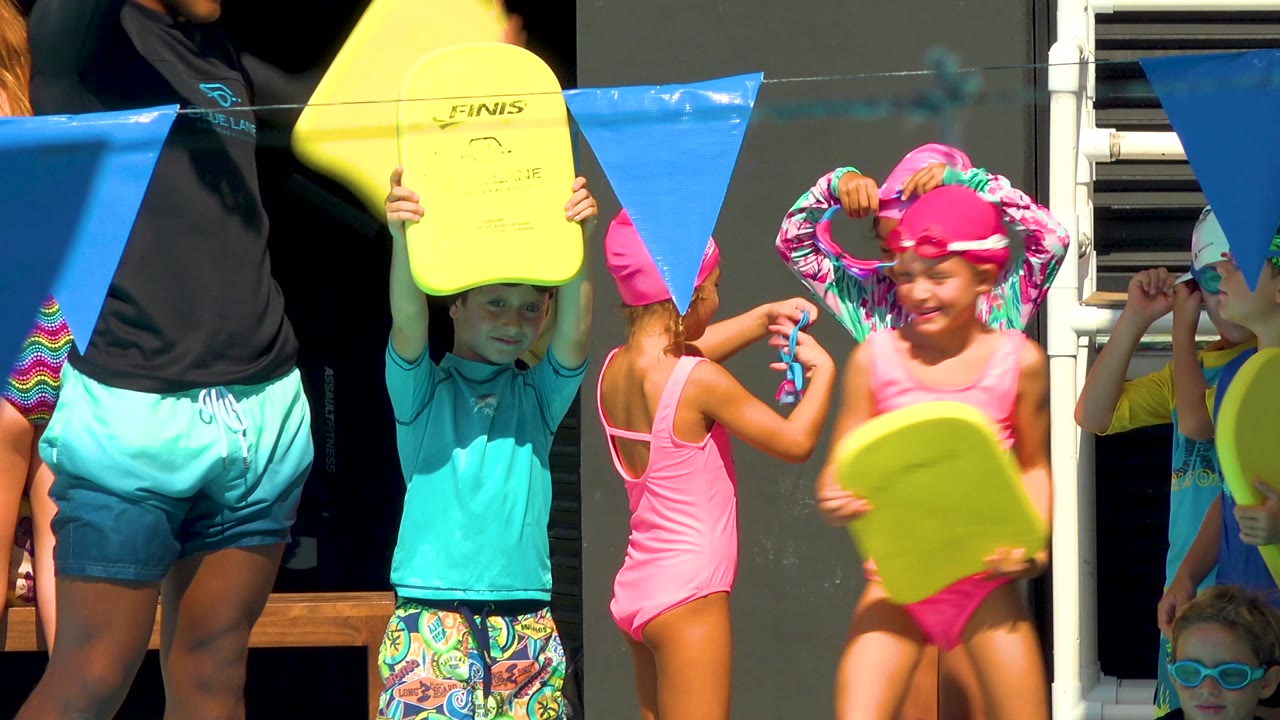 Children in Cabo San Lucas stand close together in hard sunlight, with one child holding a yellow board overhead and another adjusting a pink swim cap beneath blue flags.