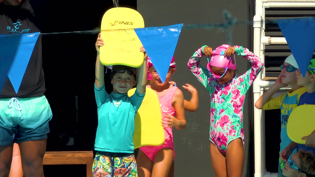 A child in a turquoise rash guard faces forward with a yellow board while an adult coach and other children in caps and swimsuits stand around blue pennant flags in Cabo San Lucas.