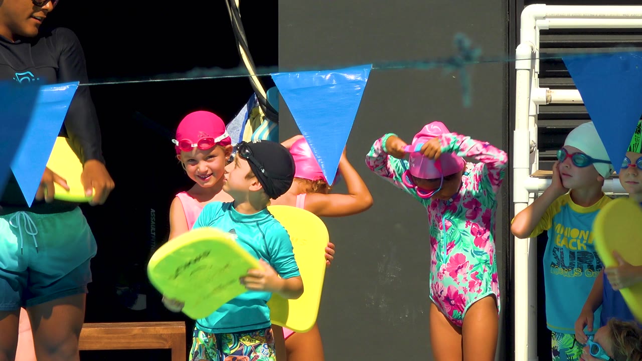 Coach holds a yellow swim board beside children in caps.