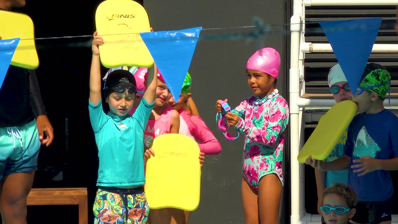 At a Cabo San Lucas pool, children in swim caps and goggles stand with yellow kickboards under blue lane pennants as an adult is partly visible at the edge.