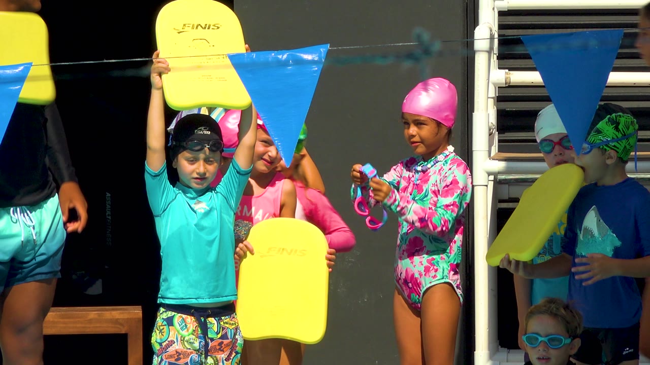 Children at the Cabo San Lucas pool cluster near a ladder and blue pennants, with one child holding a yellow kickboard high and others wearing goggles in the shade and sun.