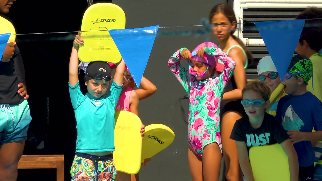 At the Cabo San Lucas pool, a girl in a bright patterned rash guard adjusts her pink cap while other children hold yellow kickboards beneath blue lane flags.