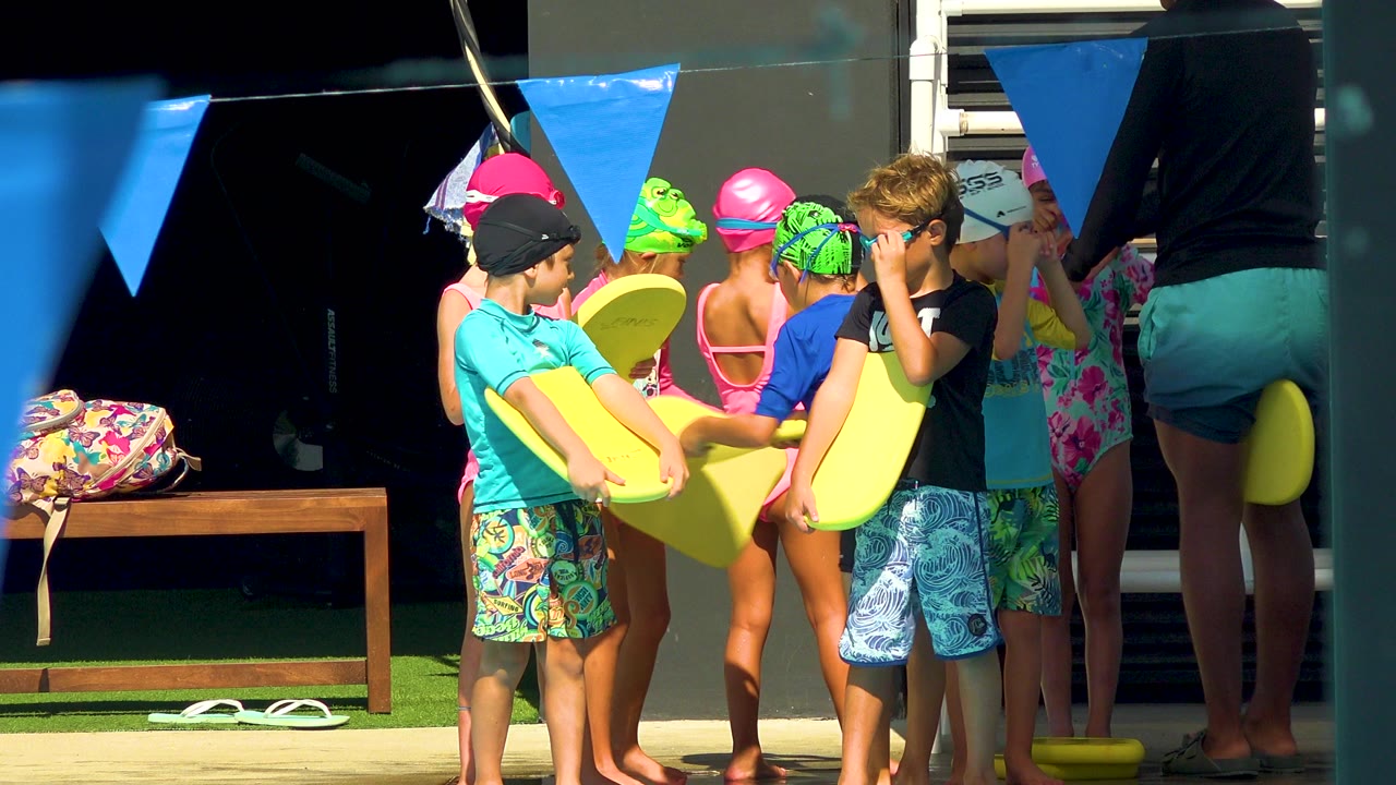 Children in swim caps and swimwear hold yellow surf planks near a shaded doorway in Cabo San Lucas, Mexico, with blue pennant flags overhead.