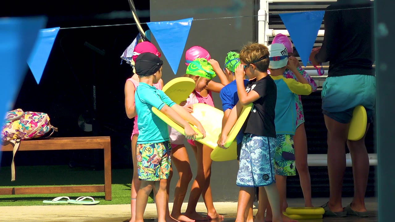 Children wearing bright caps and goggles hold yellow surf planks while standing by blue flags and a dark shaded doorway in Cabo San Lucas, Mexico.